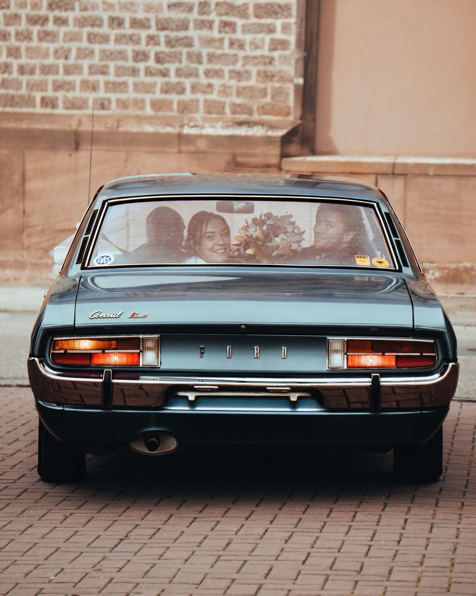 Brautpaar blickt durch die Heckscheibe eines blauen Oldtimers